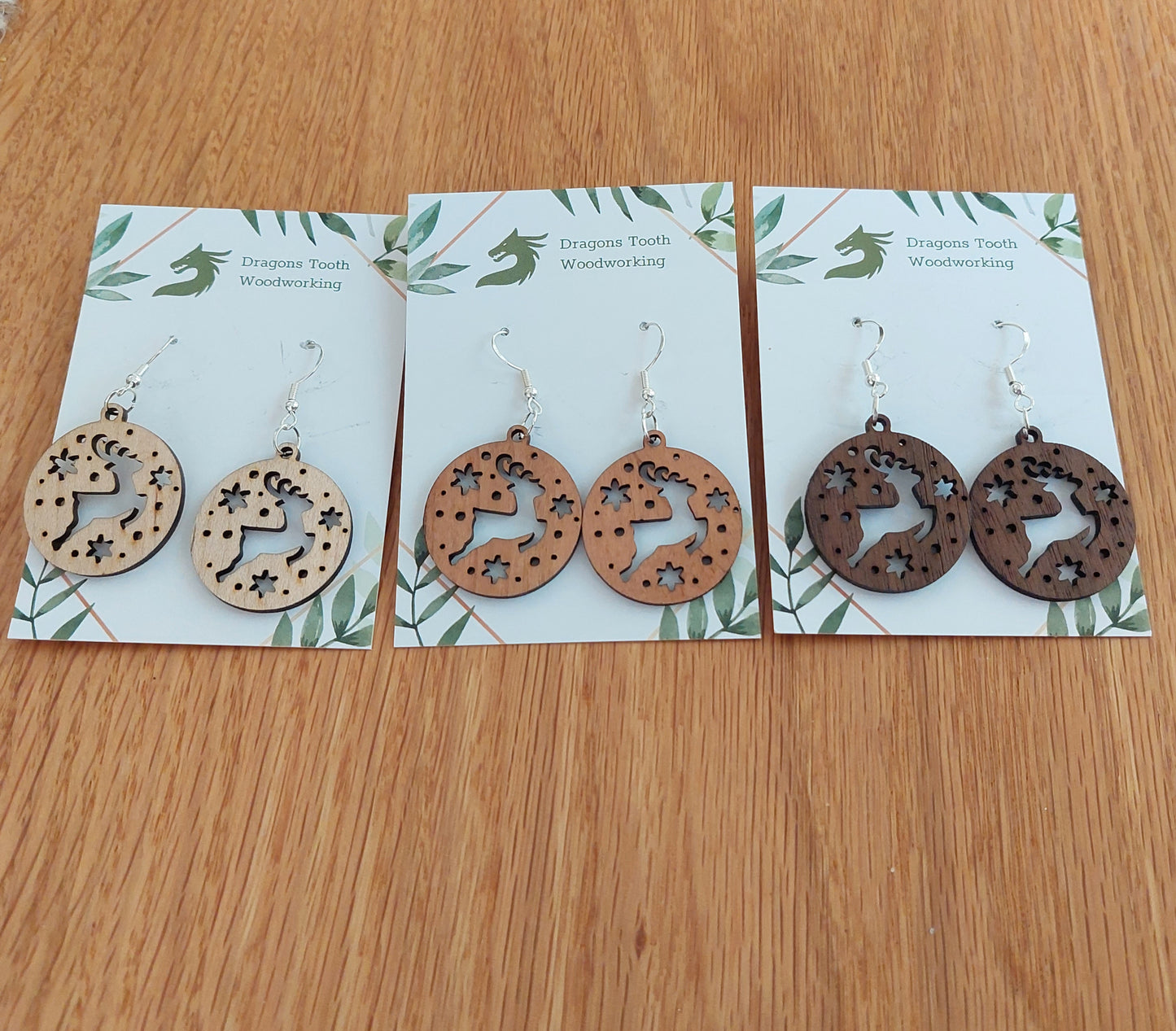 Pairs of reindeer earrings made from natural hardwoods, with silver hooks displayed on a card with 'Dragon's Tooth Woodworking' written at the top.