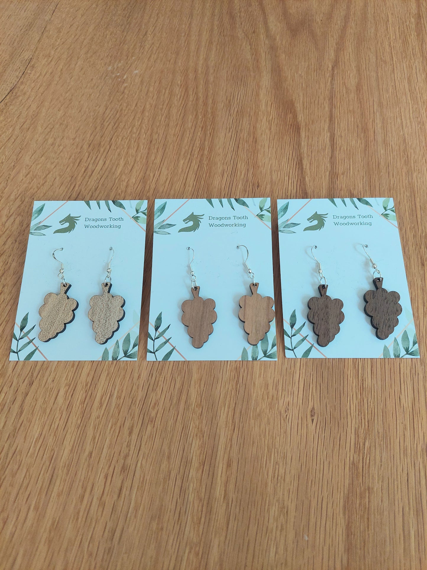 An alternate view displaying the grape cluster earrings in three available wood types: light Maple, warm Cherry, and deep Walnut.