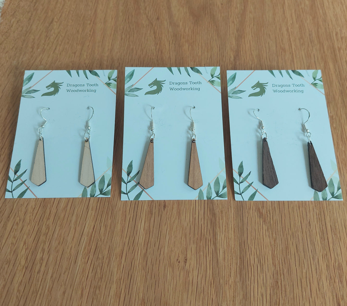 An alternate view displaying the tie silhouette geometric dangle earrings in three available wood types: light Maple, warm Cherry, and deep Walnut.