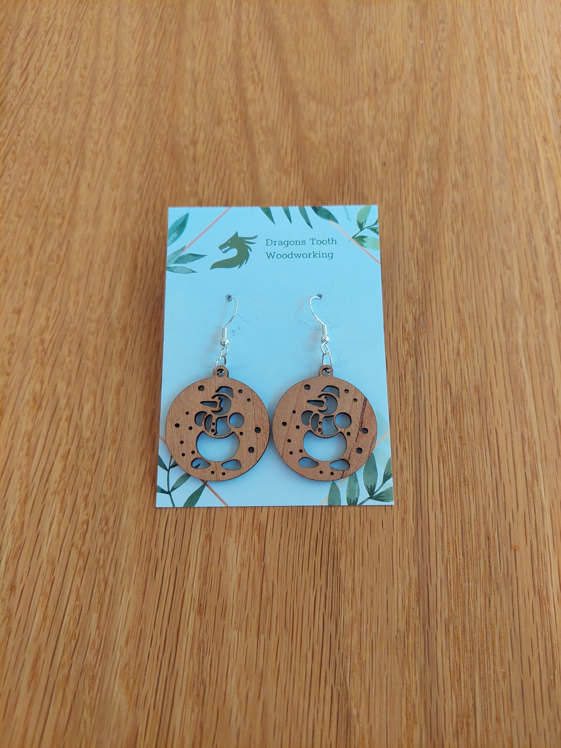A pair of snowman-shaped earrings made from natural cherry wood, with silver hooks displayed on a card