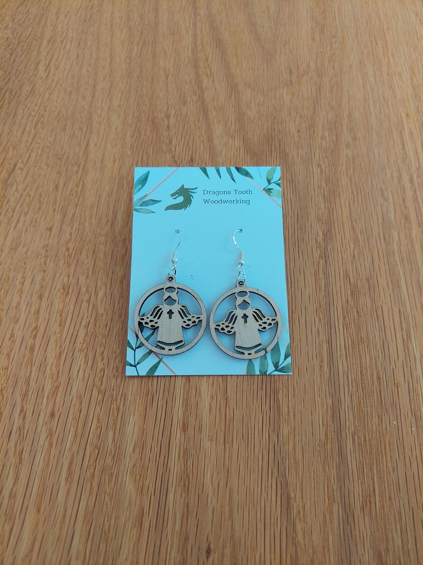 A pair of light blonde colored maple wood angel earrings displayed with silver hooks on a card with the brand name 'Dragon's Tooth Woodworking'.