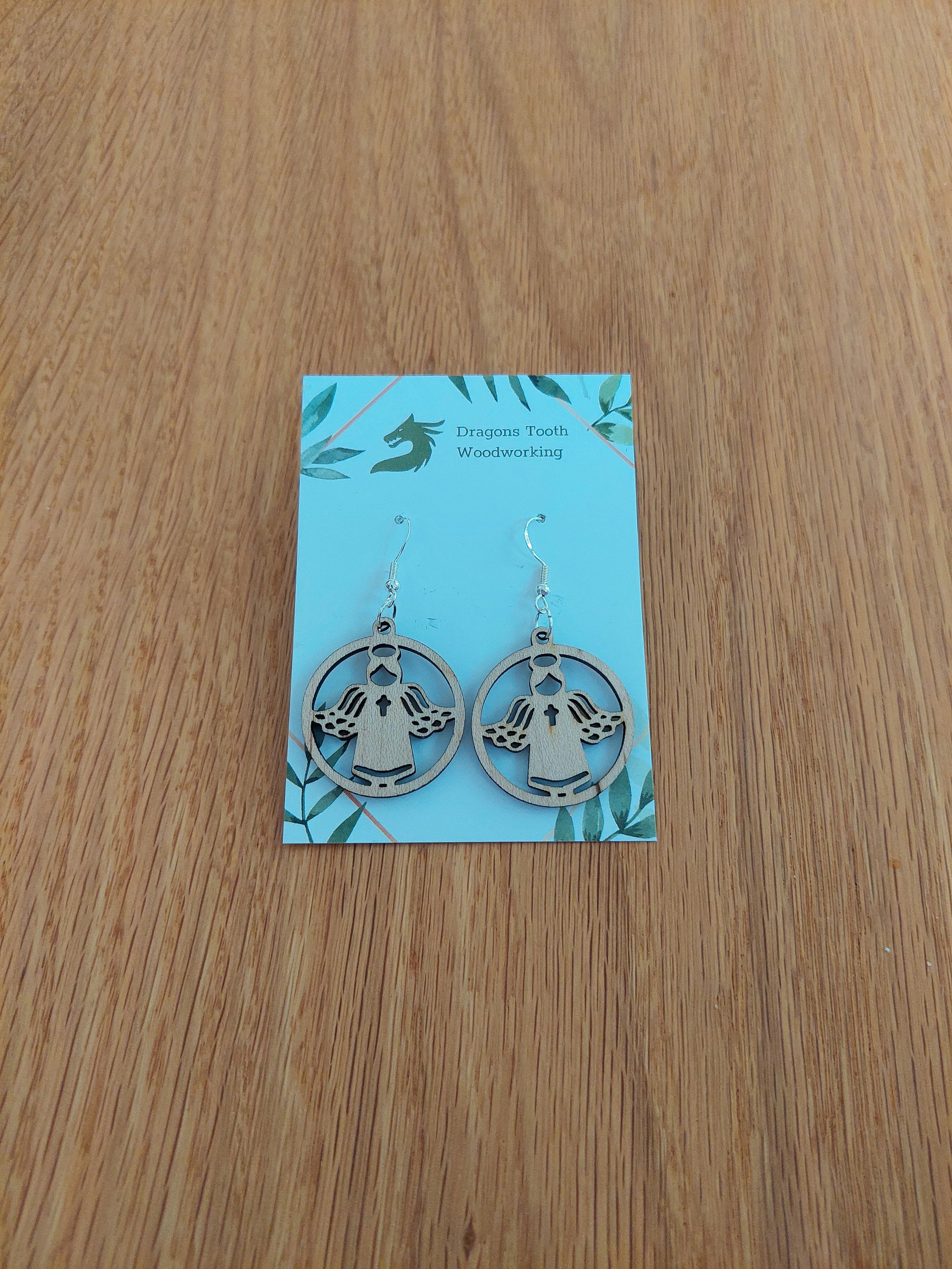 A pair of light blonde colored maple wood angel earrings displayed with silver hooks on a card with the brand name 'Dragon's Tooth Woodworking'.