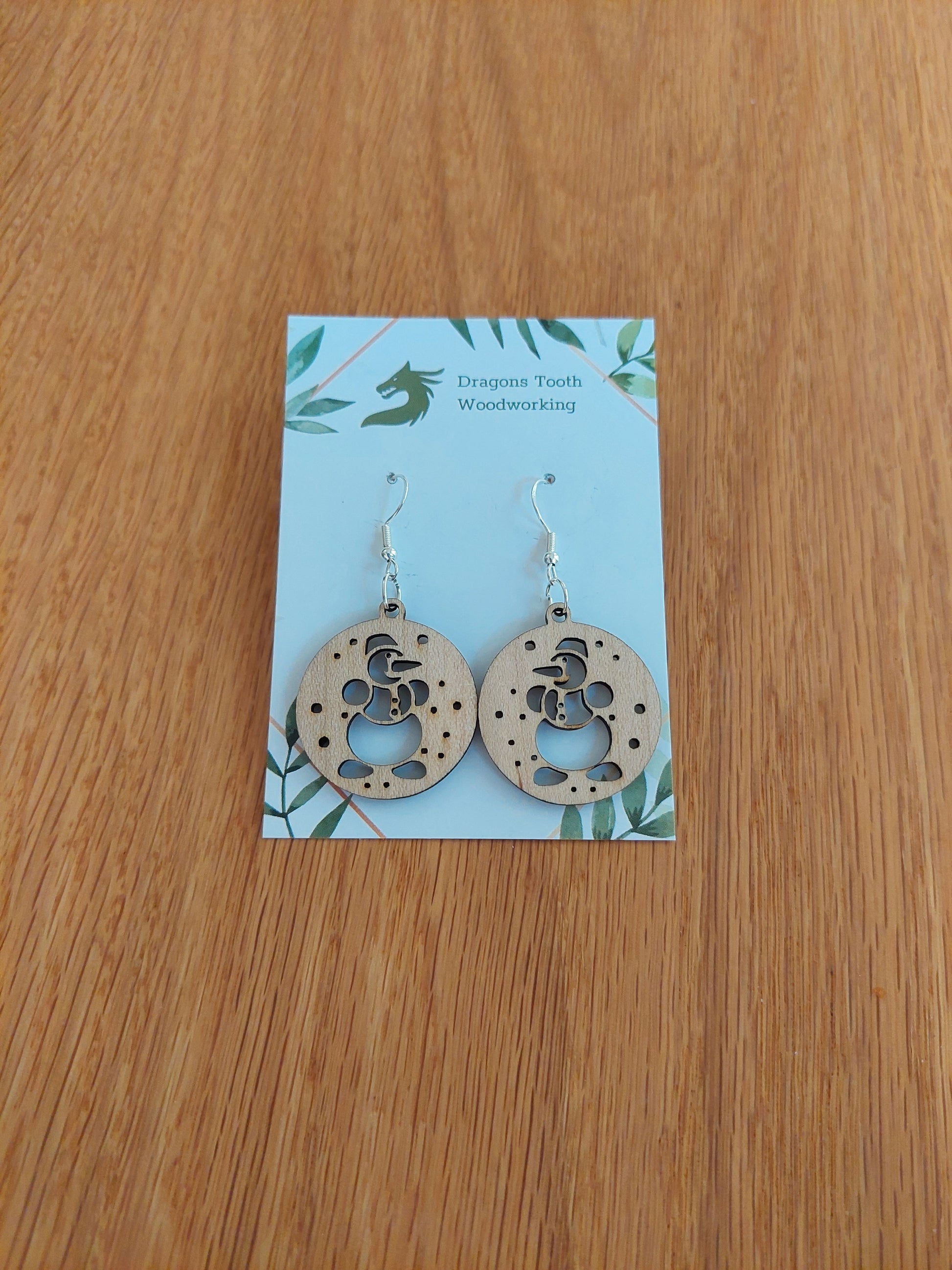 A pair of snowman-shaped earrings made from natural maple, displayed on a card