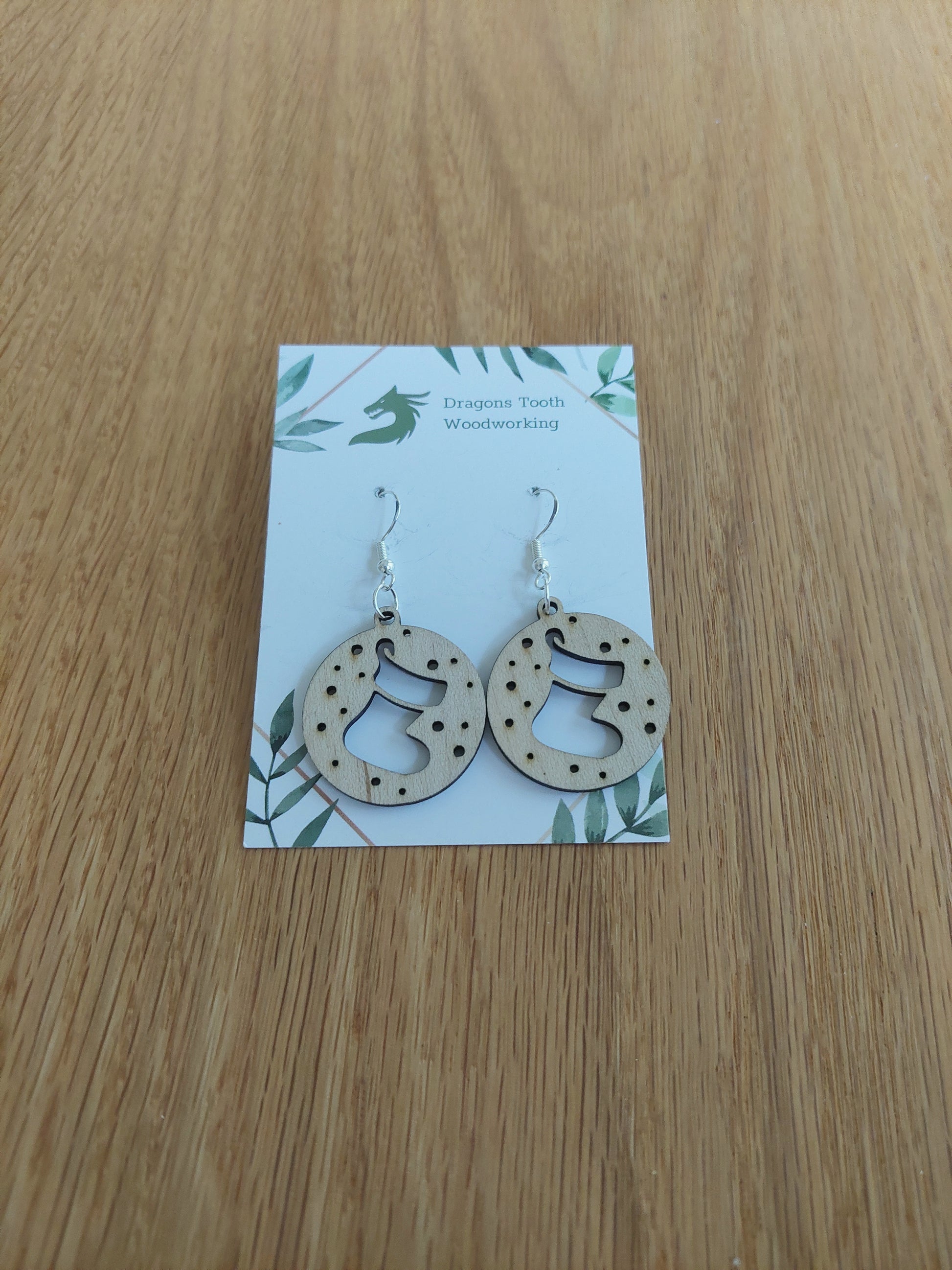 A pair of natural wood earrings featuring Christmas Stockings with hypoallergenic sterling silver plated hooks, displayed on a white card with 'Dragon's Tooth Woodworking' branding.