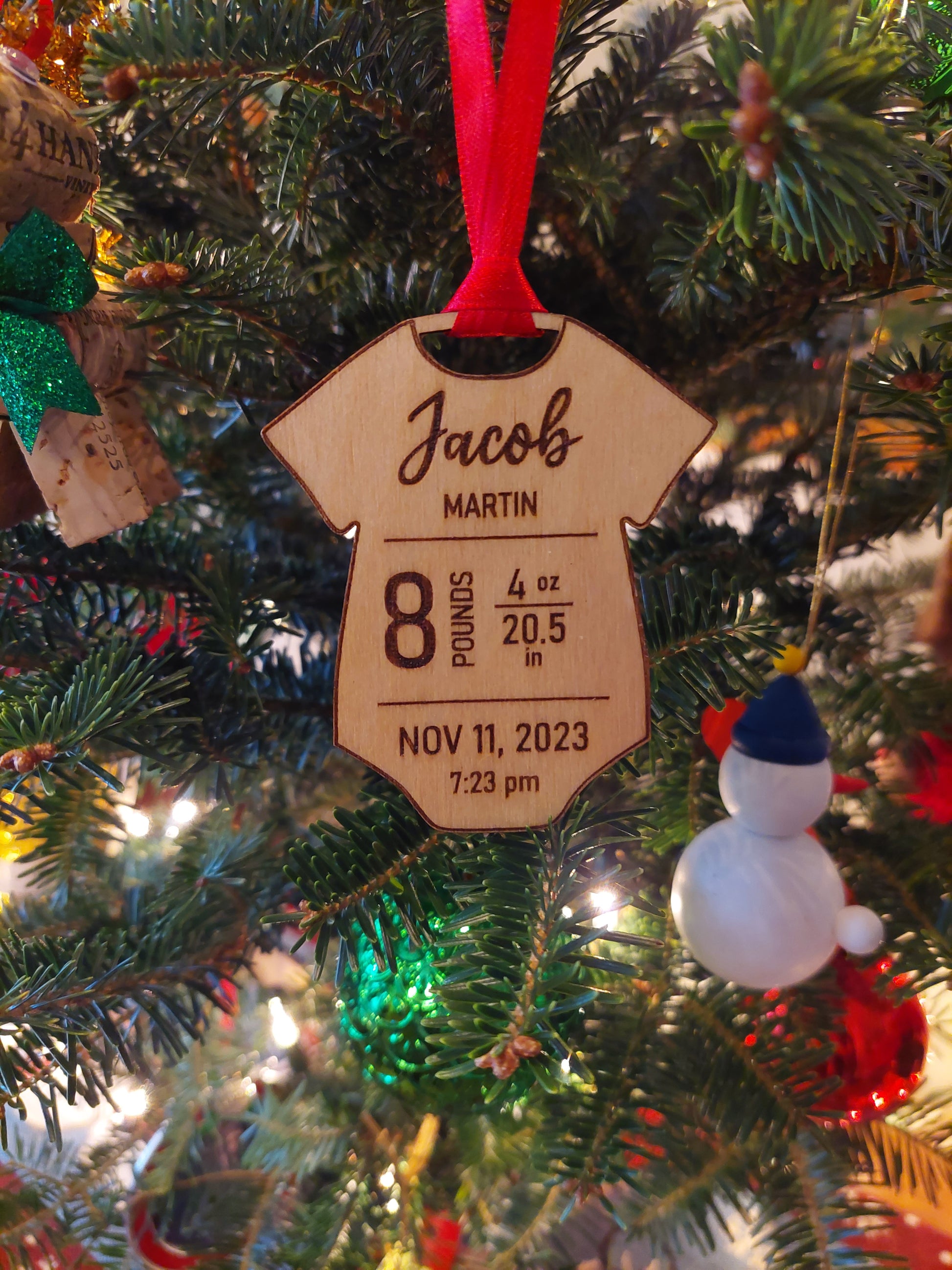 A wooden onesie-shaped ornament with engraved text displaying birth statistics, hanging on a Christmas tree with festive decorations.