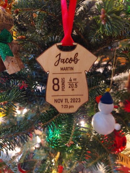 A wooden onesie-shaped ornament with engraved text displaying birth statistics, hanging on a Christmas tree with festive decorations.