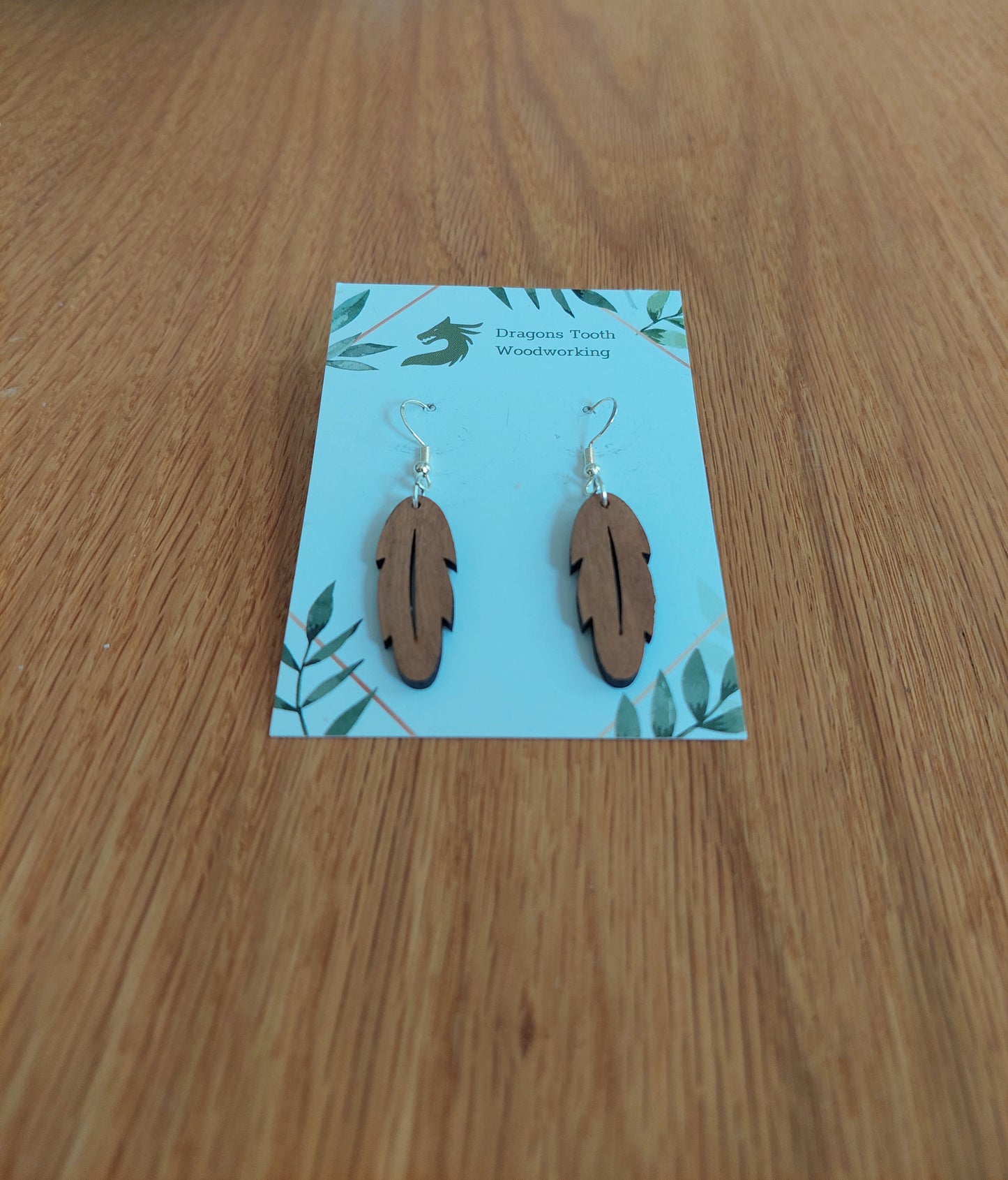 A front view of the intricate feather silhouette earrings crafted from warm, traditional caramel-toned solid Cherry wood, displayed on an earring card.