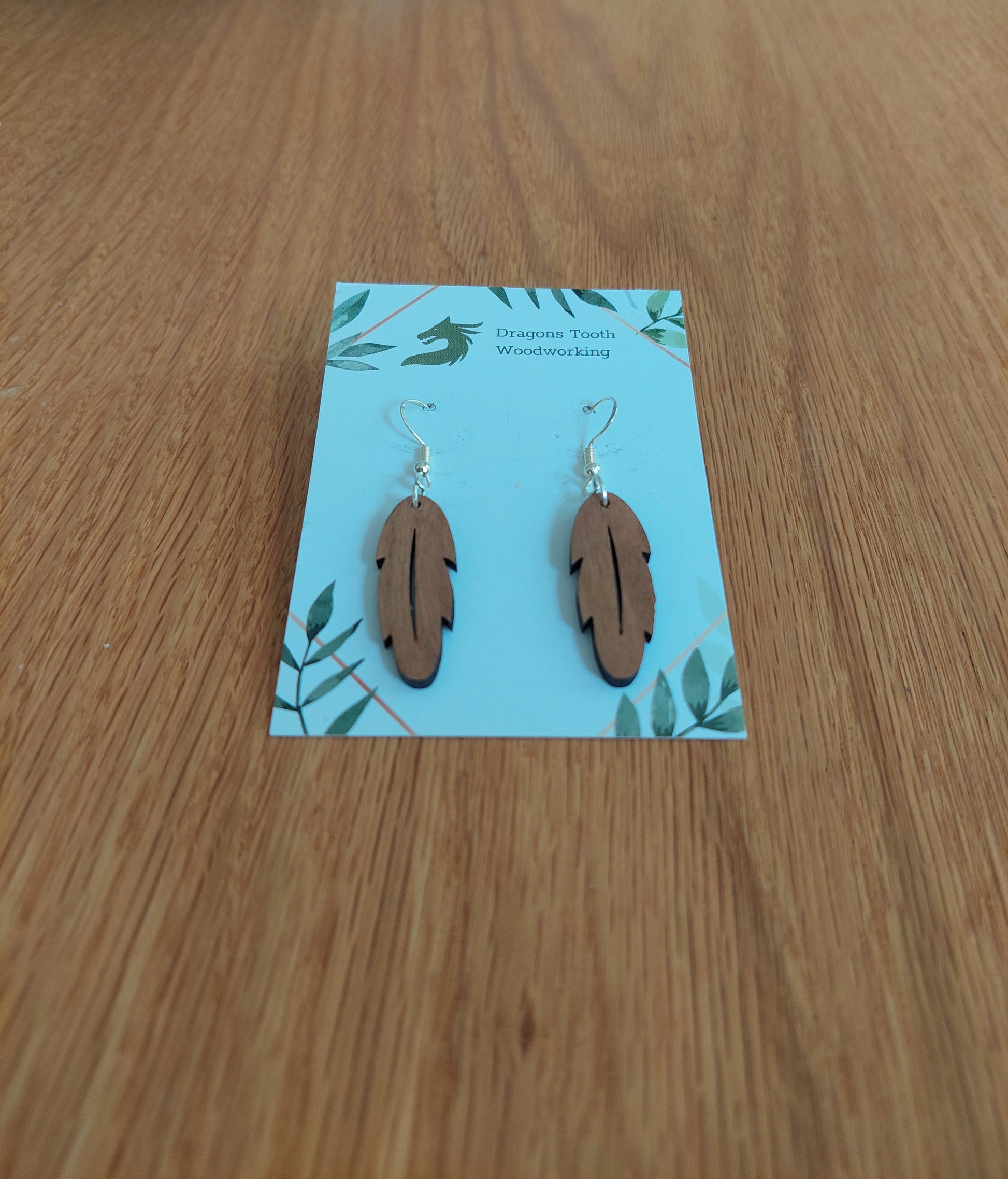 A front view of the intricate feather silhouette earrings crafted from warm, traditional caramel-toned solid Cherry wood, displayed on an earring card.