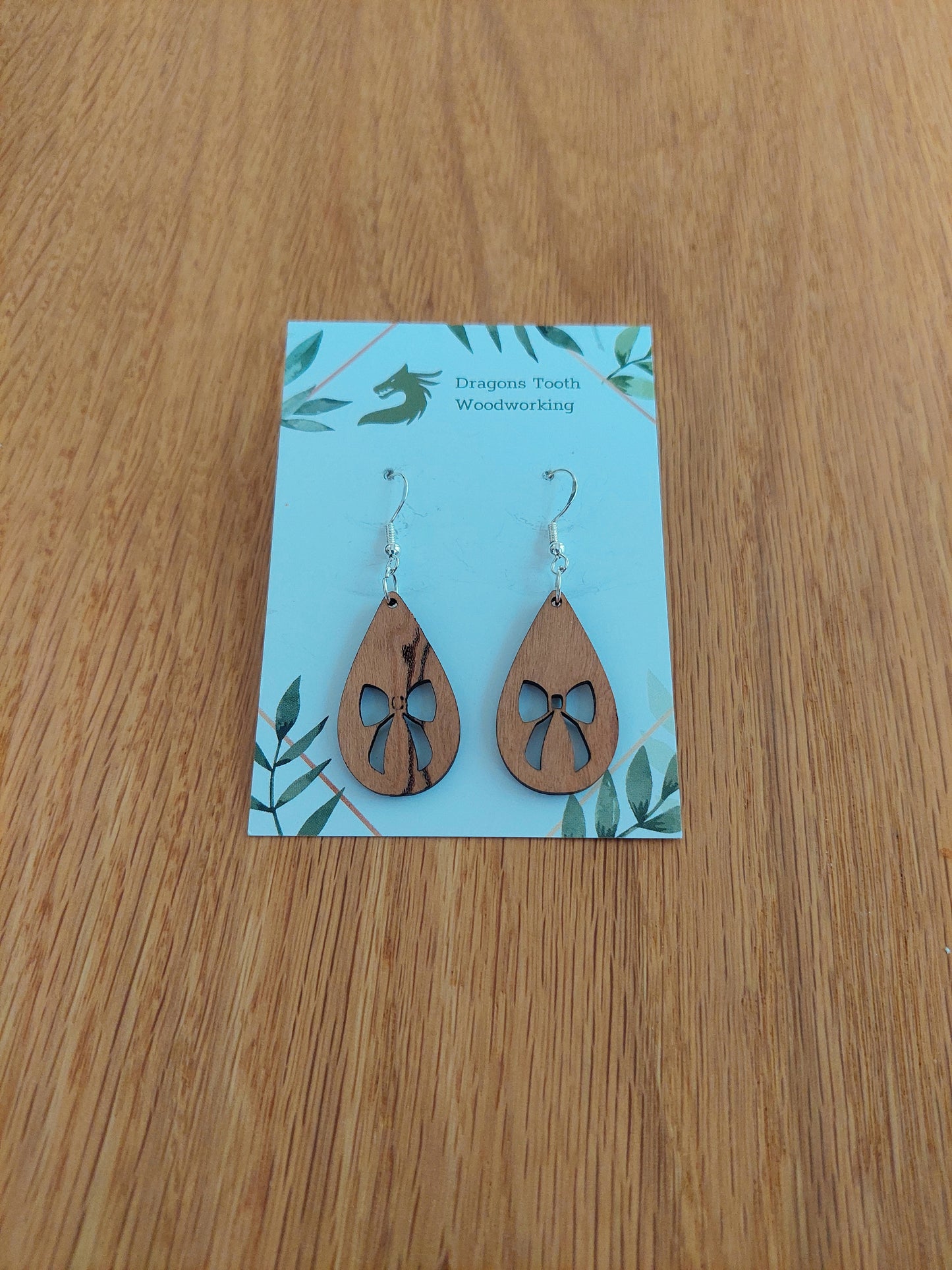 A front view of the ribbon bow teardrop earrings crafted from warm, traditional caramel-toned solid Cherry wood, displayed on an earring card.