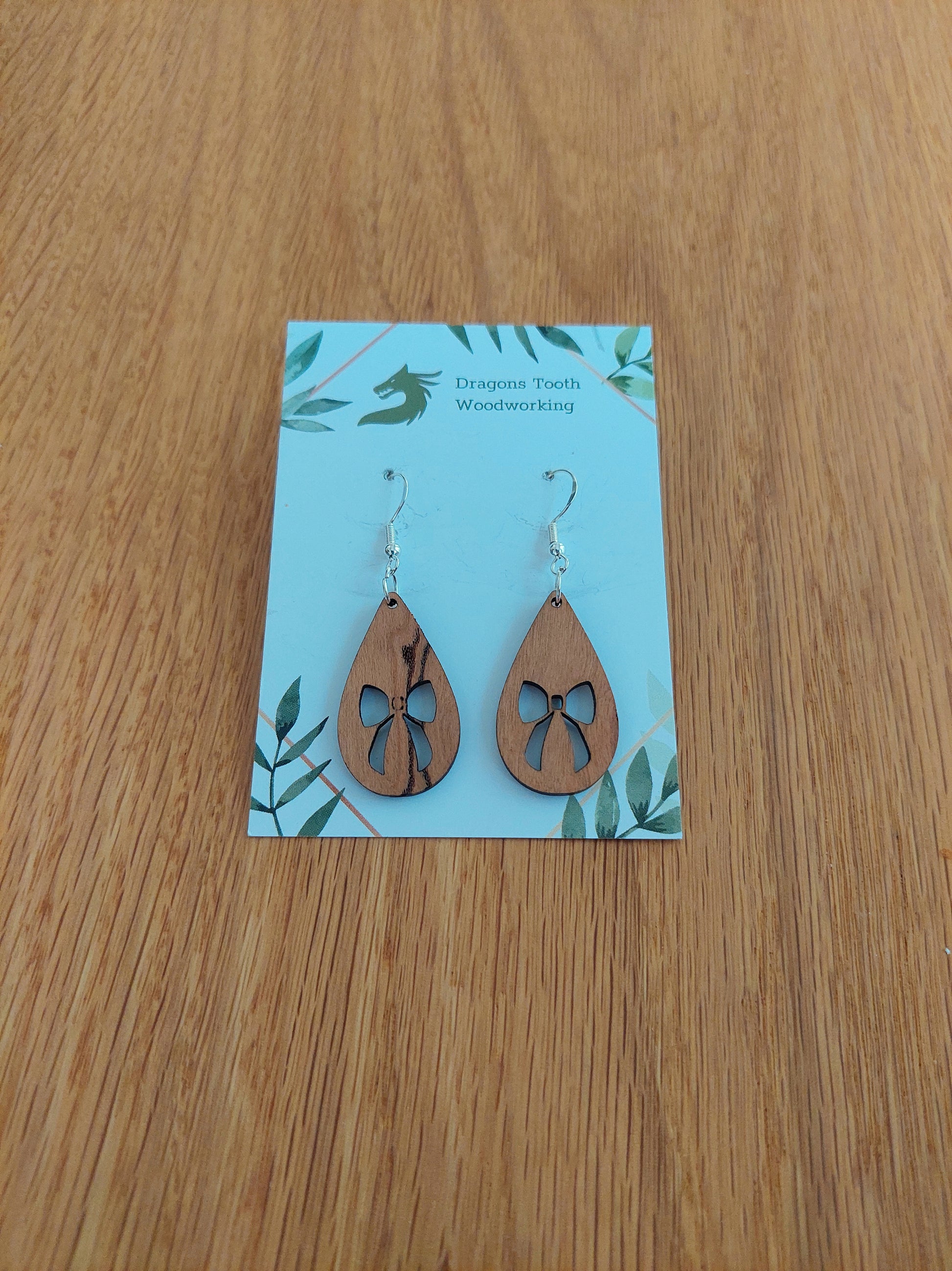 A front view of the ribbon bow teardrop earrings crafted from warm, traditional caramel-toned solid Cherry wood, displayed on an earring card.