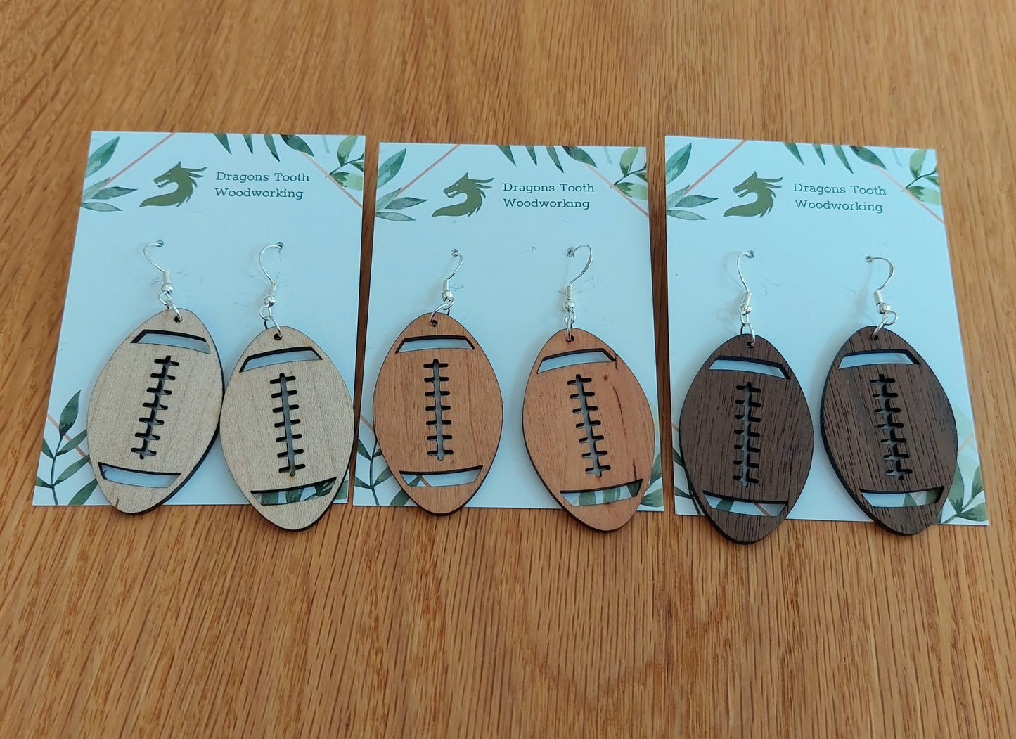 A lineup of handcrafted wooden football dangle earrings displayed together in natural walnut, cherry, and maple hardwood finishes.