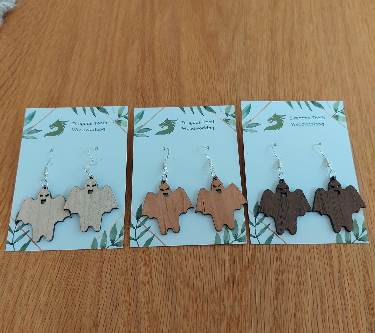 A collection of handcrafted wooden ghoul ghost dangle earrings displayed in natural maple, cherry, and walnut hardwood finishes.