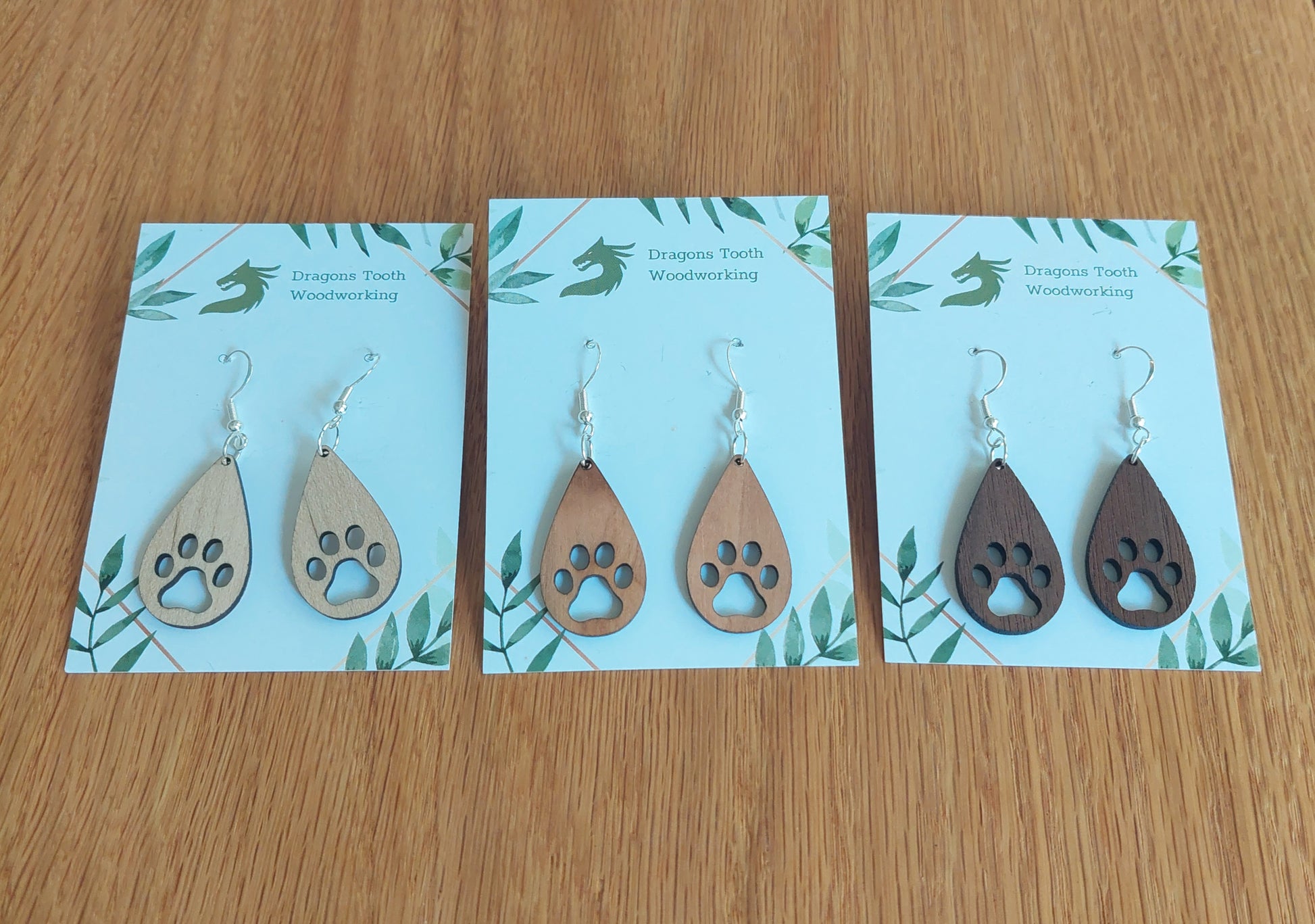 A collection of handcrafted wooden pawprint dangle earrings displayed together in natural maple, cherry, and walnut wood finishes.