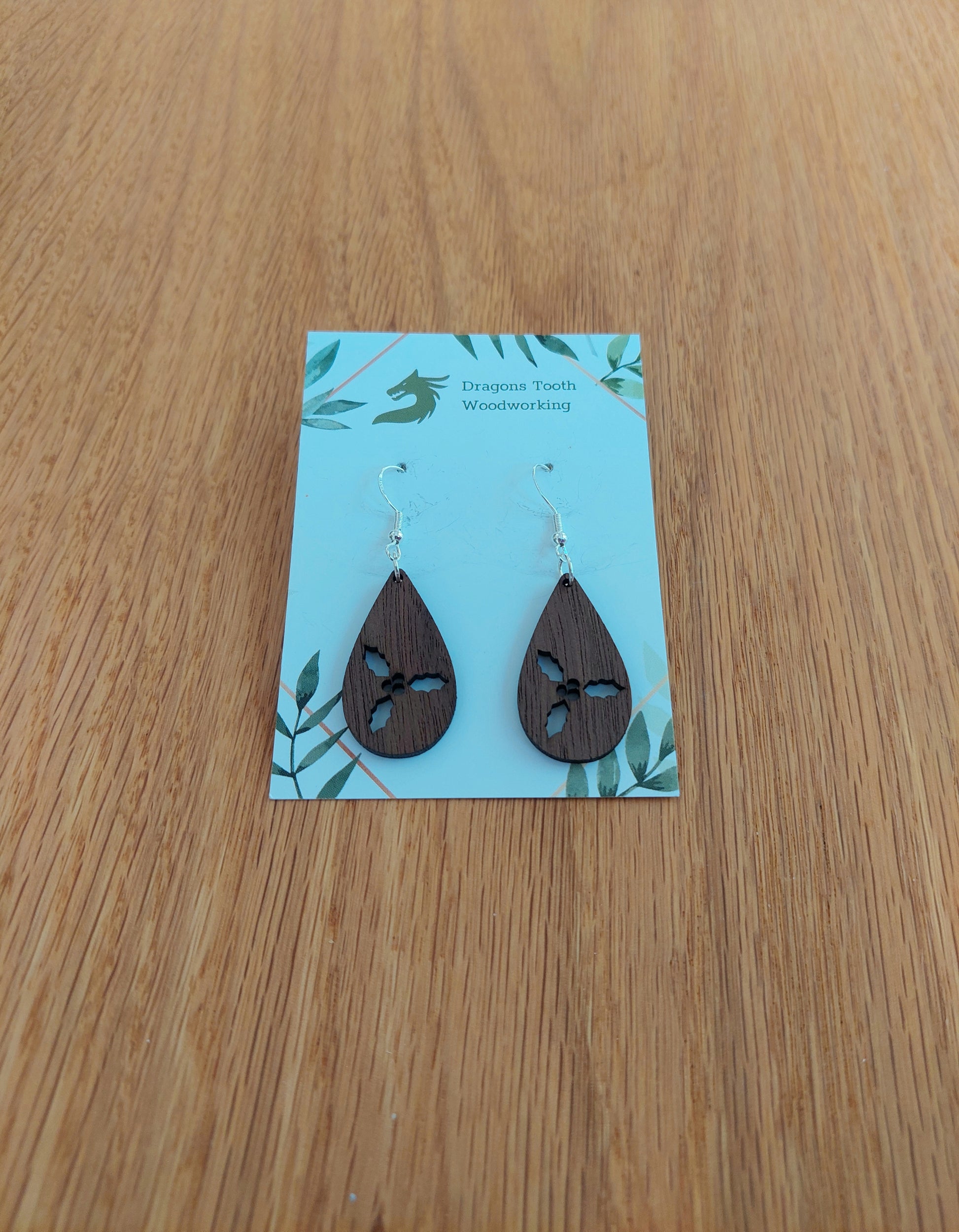 A pair of teardrop shaped earrings made from natural walnut hardwood, displayed on a card. The earring is laser-cut with a holly cluster design.
