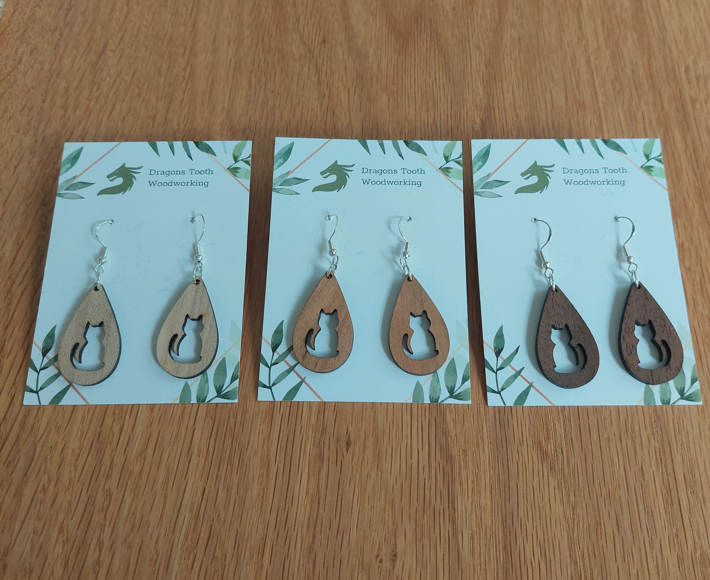 An alternate view displaying the circular cat earrings in three available wood types: light Maple, warm Cherry, and deep Walnut.