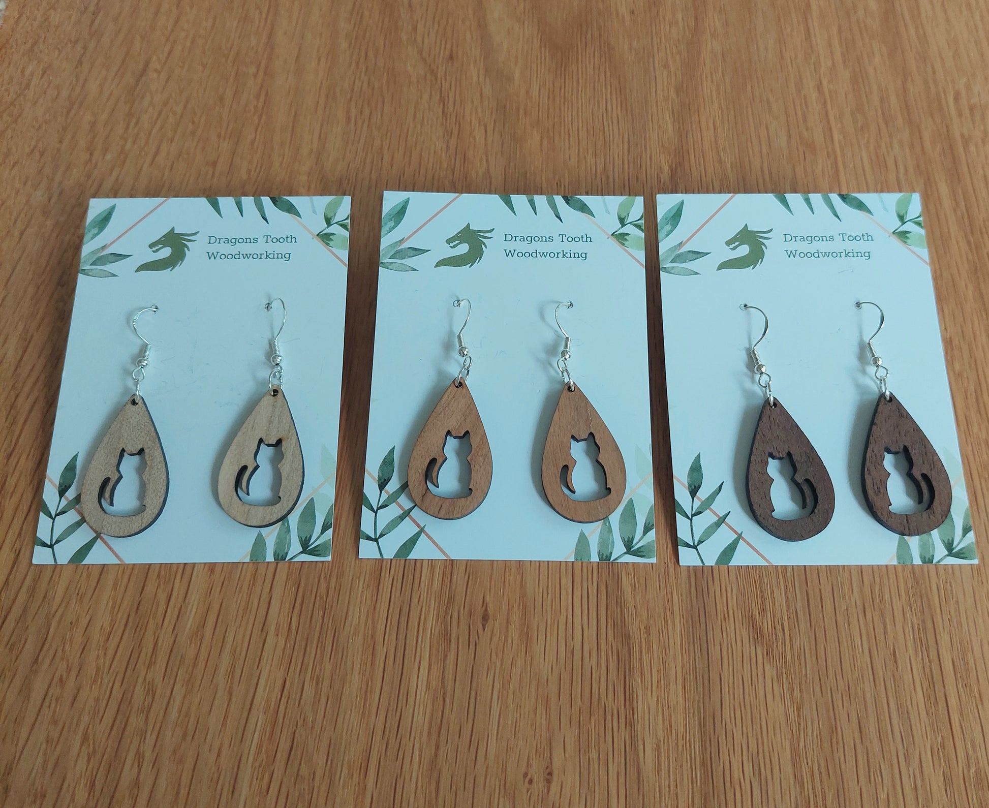 An alternate view displaying the circular cat earrings in three available wood types: light Maple, warm Cherry, and deep Walnut.