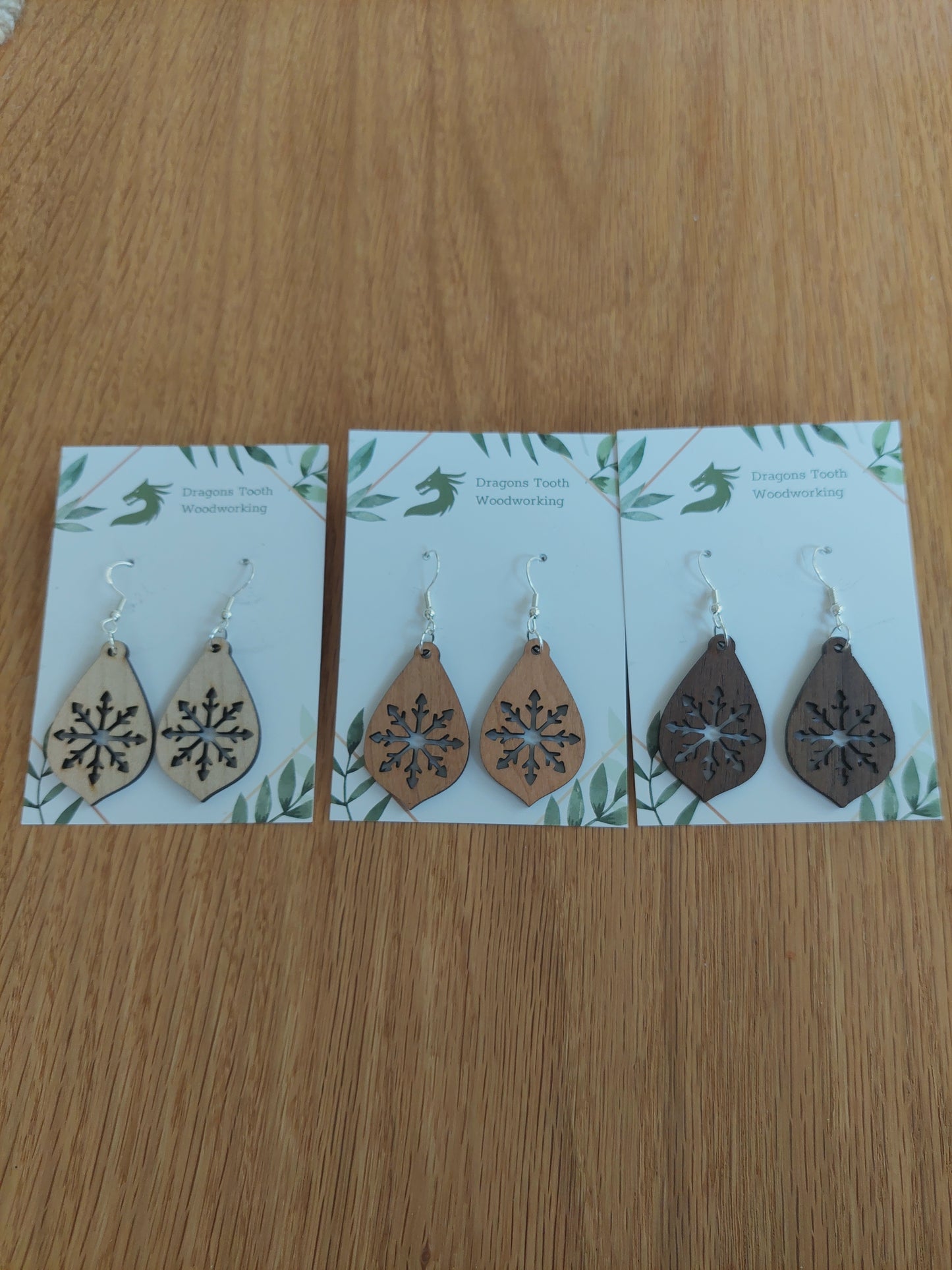 An alternate view displaying the large snowflake teardrop earrings in three available wood types: light Maple, warm Cherry, and deep Walnut.