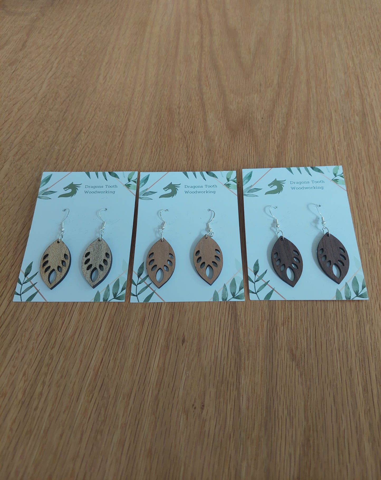An alternate view displaying the monstera leaf earrings in three available wood types: light Maple, warm Cherry, and deep Walnut.