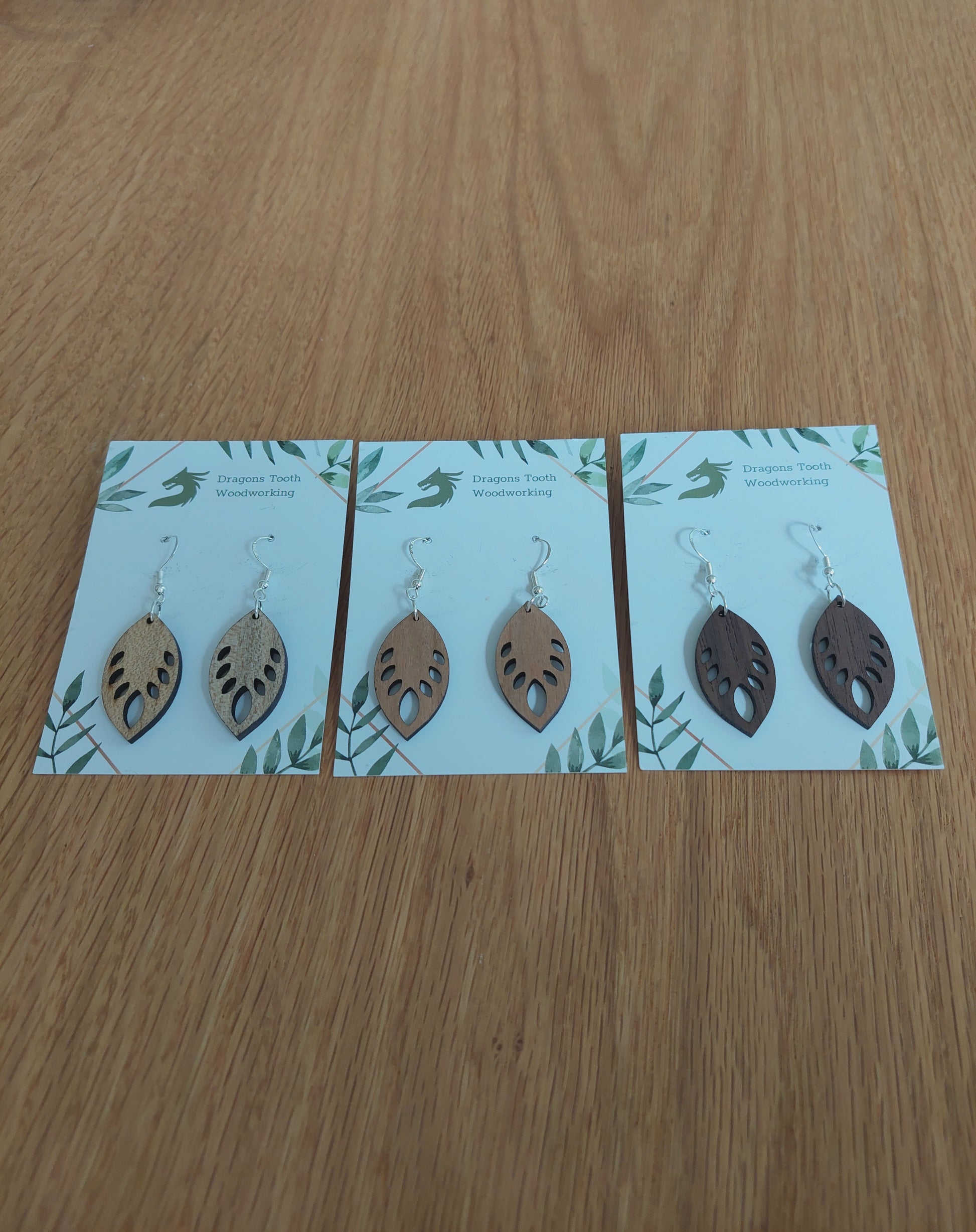An alternate view displaying the monstera leaf earrings in three available wood types: light Maple, warm Cherry, and deep Walnut.
