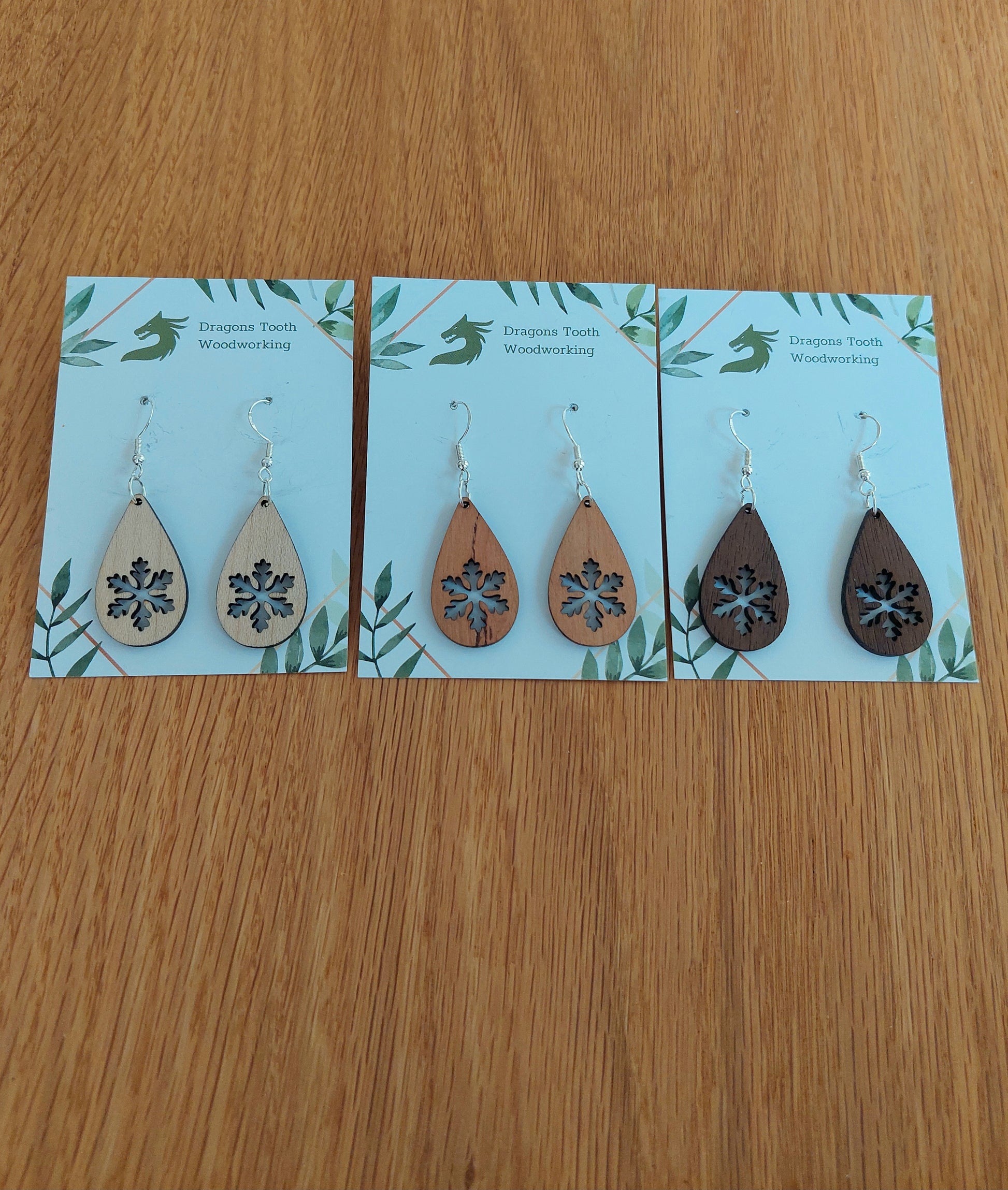 An alternate view displaying the snowflake teardrop earrings in three available wood types: light Maple, warm Cherry, and deep Walnut.