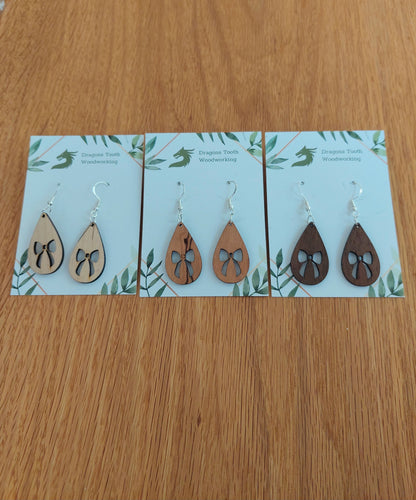 An alternate view displaying the ribbon bow teardrop earrings in three available wood types: light Maple, warm Cherry, and deep Walnut.