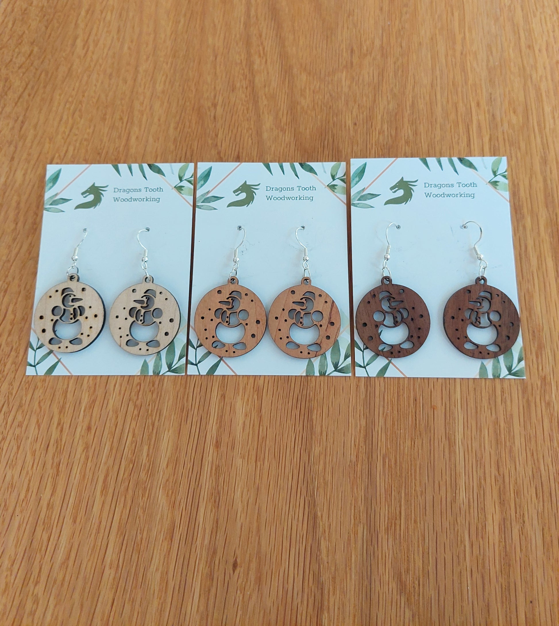 n alternate view displaying the snowman earrings in three available wood types: light Maple, warm Cherry, and deep Walnut.