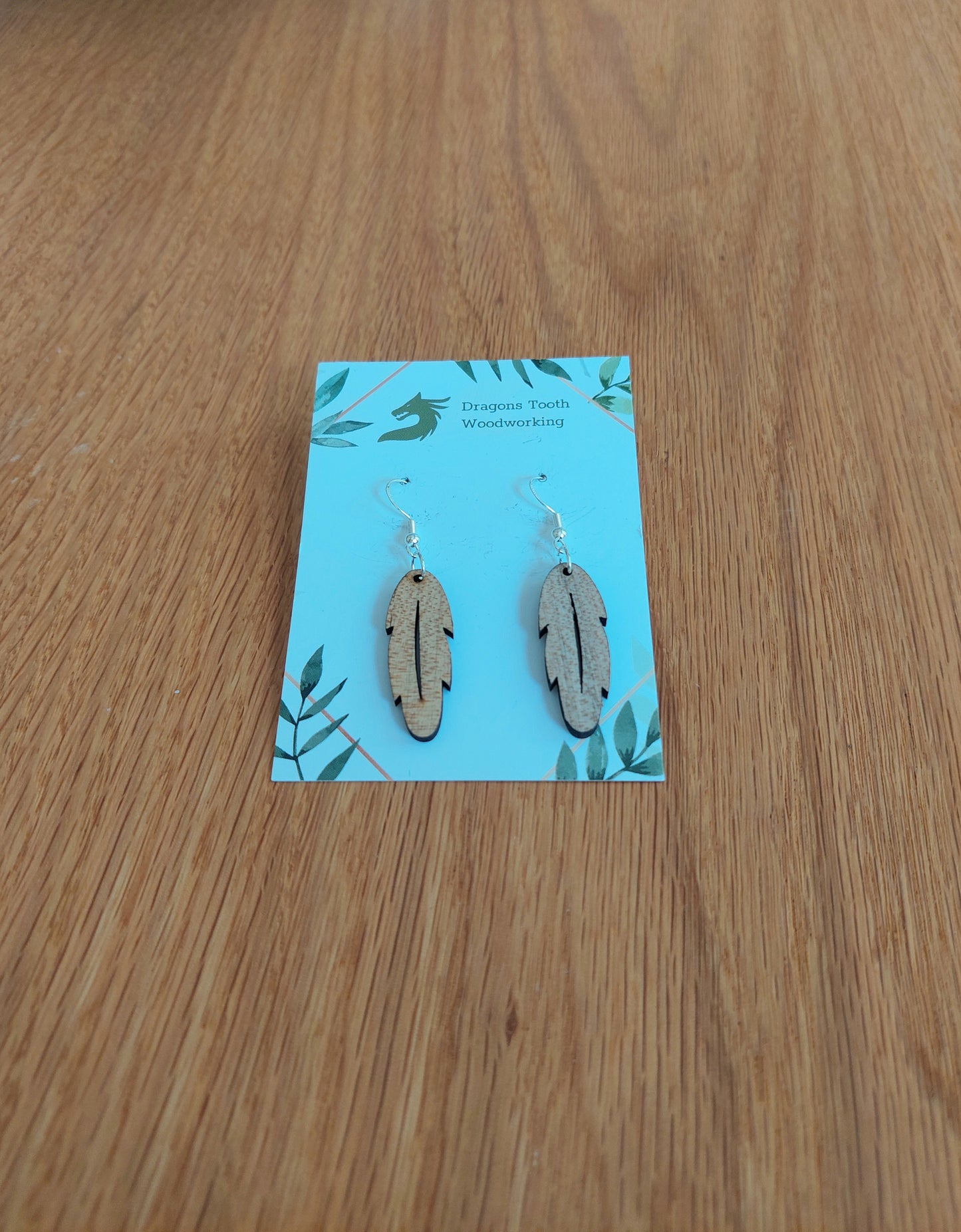 A front view of the intricate feather silhouette earrings crafted from light, bright blonde solid Maple wood, displayed on an earring card.