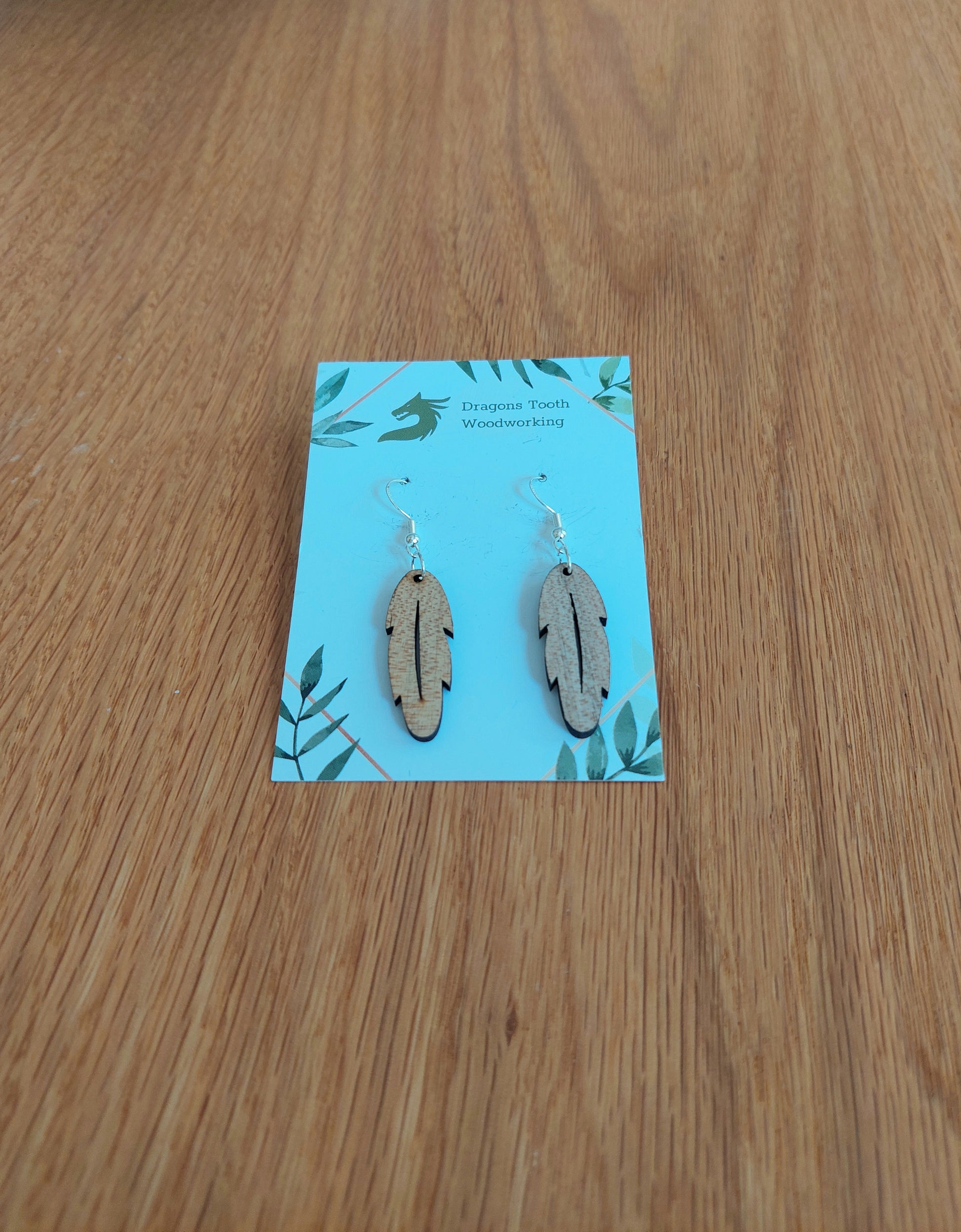 A front view of the intricate feather silhouette earrings crafted from light, bright blonde solid Maple wood, displayed on an earring card.