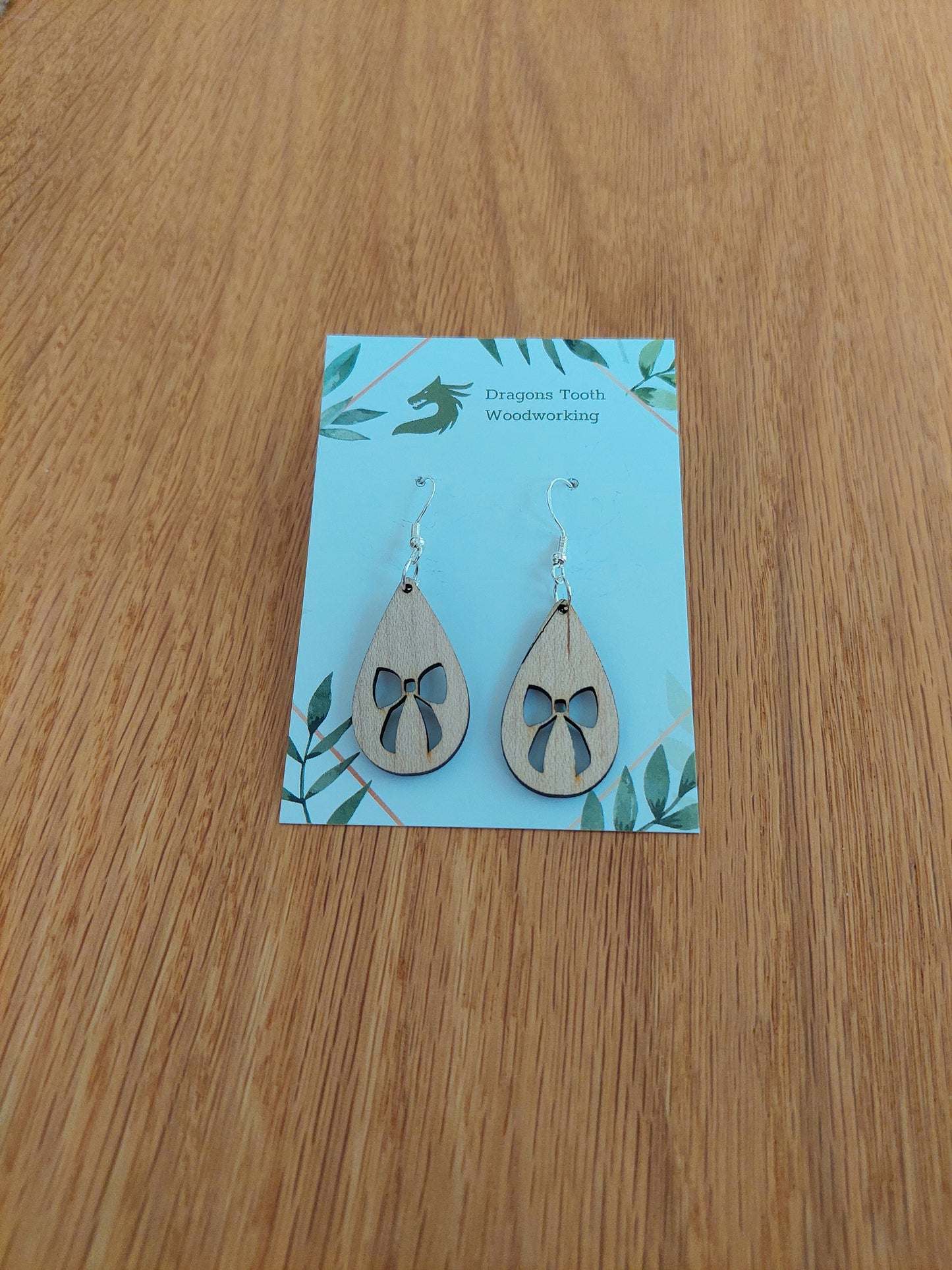 A front view of the ribbon bow teardrop earrings crafted from light, bright blonde solid Maple wood, displayed on an earring card.