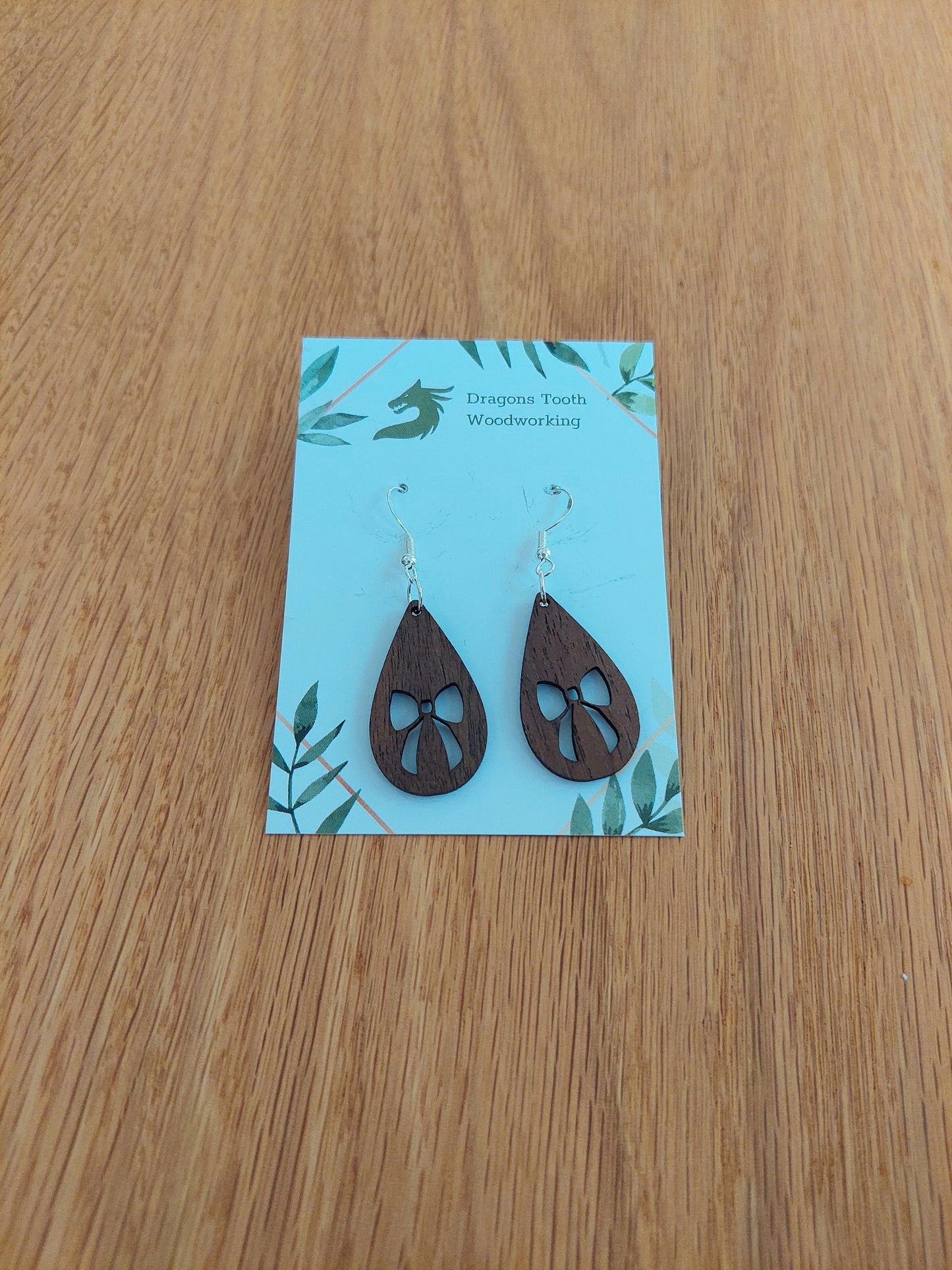 A front view of the ribbon bow teardrop earrings crafted from rich, deep chocolate-brown solid Walnut wood, displayed on an earring card.